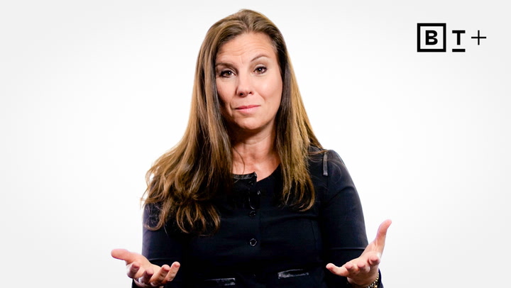 A woman with long brown hair, wearing a black top, sits facing the camera with her hands open. A "B T +" logo appears in the top right corner.