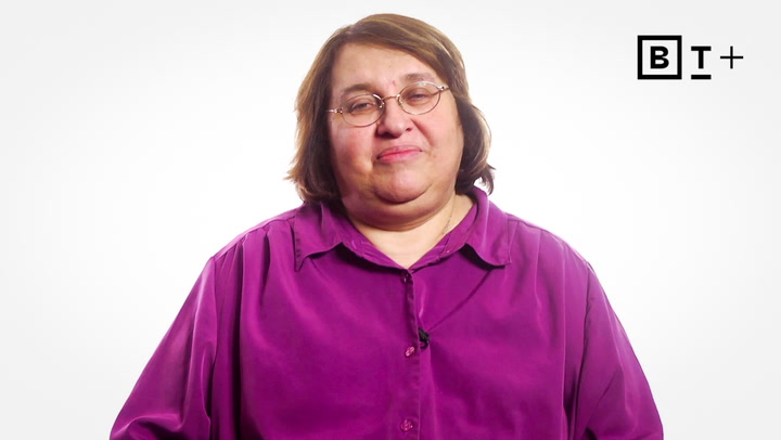 A person wearing glasses and a purple shirt is standing in front of a white background with a "B T +" logo in the top right corner.