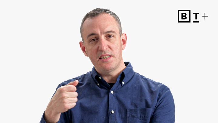 A man in a blue button-up shirt gestures with his right hand against a plain white background. A "BT+" logo is visible in the top right corner.