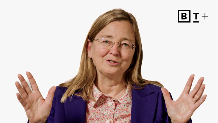 A woman with long hair and glasses speaks while gesturing with both hands, wearing a purple blazer and floral shirt, with a "B T +" logo in the top right corner.