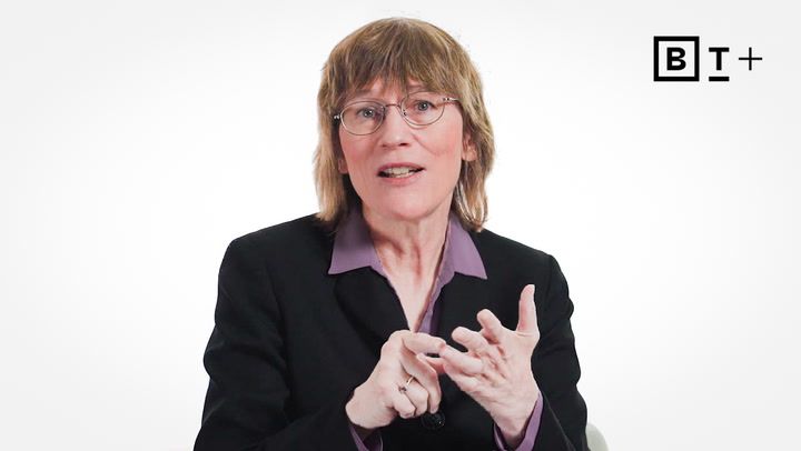 A person with glasses and shoulder-length hair, wearing a black blazer and purple shirt, gestures with their hands against a plain white background with a "BT+" logo in the corner.
