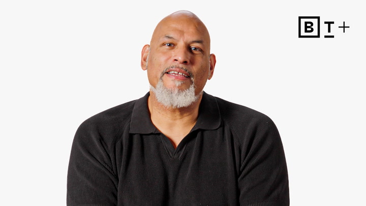 A man with a bald head and gray goatee, wearing a black collared shirt, sits in front of a plain white background with the "B T +" logo in the top right corner.