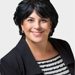 Woman with short dark hair wearing a black blazer and a black-and-white striped top, smiling at the camera against a plain white background.