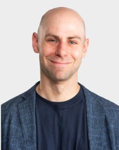 A bald man in a blue blazer and navy shirt smiles at the camera against a plain light background.