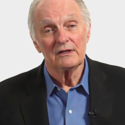 An older man with gray hair wearing a black suit jacket and blue shirt faces the camera with a neutral expression.