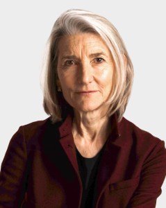 An older woman with shoulder-length gray hair, wearing a dark burgundy jacket over a black top, looks directly at the camera with a neutral expression against a plain background.