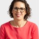 A woman with short, curly brown hair and glasses is wearing a coral top and a necklace, smiling at the camera against a plain light background.
