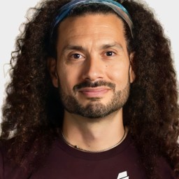 A person with long curly hair, a beard, and a headband, wearing a dark shirt that reads "everyday spy" on a plain background.