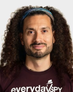 A person with long curly hair, a beard, and a headband, wearing a dark shirt that reads "everyday spy" on a plain background.
