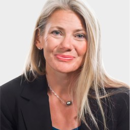 A woman with long blonde hair, wearing a black blazer, blue top, and necklace, poses against a plain light background.