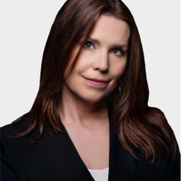 Woman with straight brown hair wearing a black blazer and white top, looking at the camera with a neutral expression against a plain light background.