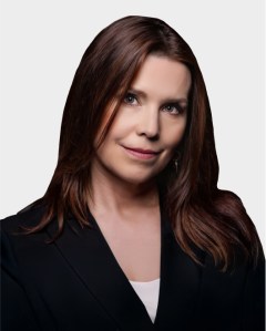 Woman with straight brown hair wearing a black blazer and white top, looking at the camera with a neutral expression against a plain light background.