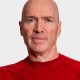 A middle-aged man with a bald head and light skin wearing a red sweater, facing forward against a plain light background.