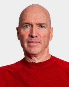 A middle-aged man with a bald head and light skin wearing a red sweater, facing forward against a plain light background.