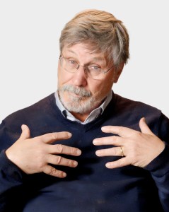 Older man with glasses and gray hair, wearing a dark sweater and collared shirt, gestures toward himself with both hands against a plain light background.