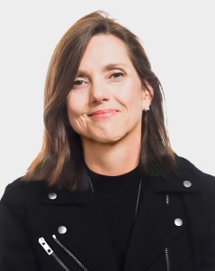 A woman with straight brown hair wearing a black jacket and a black top, smiling slightly, is posing against a plain white background.