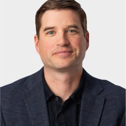 A man with short brown hair wearing a dark blazer and black shirt, facing forward against a plain light background.