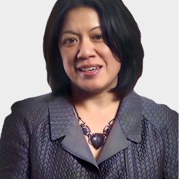 A woman with shoulder-length dark hair wears a textured gray blazer and a chunky black necklace, standing against a plain light background.