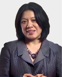 A woman with shoulder-length dark hair wears a textured gray blazer and a chunky black necklace, standing against a plain light background.