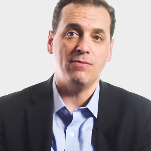 A man with short dark hair wearing a blue shirt and dark blazer poses in front of a plain light background.