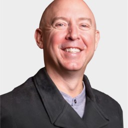 A bald man wearing a dark coat and light shirt smiles at the camera against a plain white background.