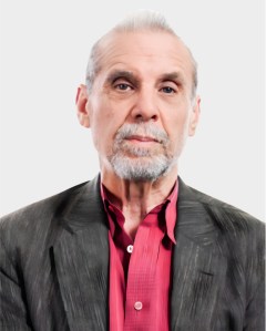 An older man with gray hair and beard wearing a dark jacket over a red shirt, looking straight at the camera against a plain light background.