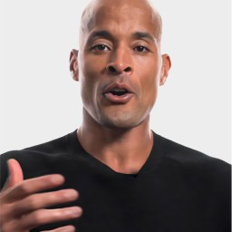 A man with a shaved head and medium skin tone, wearing a black shirt, gestures with his right hand while speaking against a plain light background.