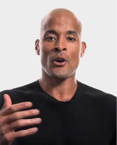 A man with a shaved head and medium skin tone, wearing a black shirt, gestures with his right hand while speaking against a plain light background.