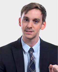 A man in a dark suit, light blue shirt, and plaid tie is facing the camera against a plain light background.