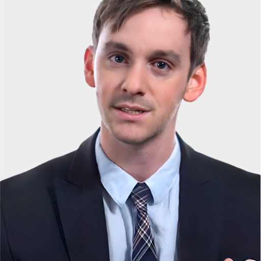 A man in a dark suit, light blue shirt, and plaid tie is facing the camera against a plain light background.