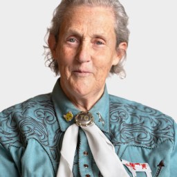 An older woman with gray hair wears a teal embroidered shirt, a white scarf with a bolo tie, and looks directly at the camera against a plain background.