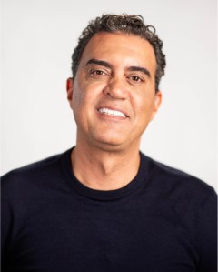 A man with short curly hair wearing a black crew-neck shirt is smiling at the camera against a plain, light background.