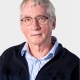 Older man with short gray hair and glasses, wearing a striped shirt and dark jacket, looking at the camera against a plain light background.
