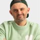 A man with a beard wearing a green sweater with a white heart and a backwards green cap, smiling with arms crossed against a plain background.