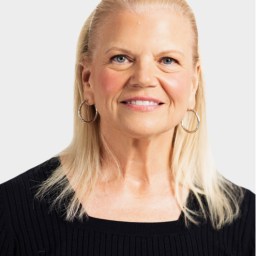 A woman with straight, shoulder-length blonde hair, wearing hoop earrings and a black ribbed top, smiles at the camera against a plain light background.