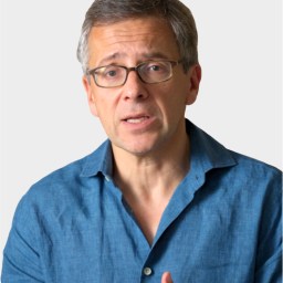 A middle-aged man with glasses and gray hair, wearing a blue shirt, speaks while gesturing with his hands against a plain background.