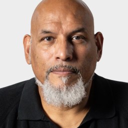 A middle-aged man with a bald head, gray goatee, and mustache, wearing a black collared shirt, facing forward against a plain background.