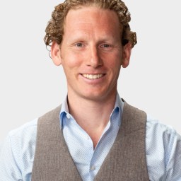 A man with curly hair, wearing a light blue dress shirt and a grey vest, smiles at the camera against a plain background.