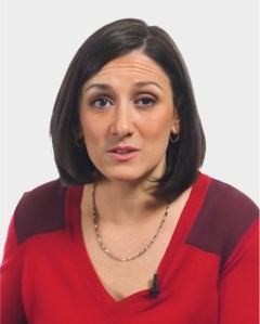 A woman with straight, shoulder-length dark hair wears a red blouse and a necklace, facing forward with a neutral expression against a plain white background.