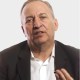 A middle-aged man in a dark suit and white shirt speaks, gesturing with his hand, against a plain light background.