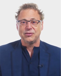 A middle-aged man with light skin, glasses, and short, curly hair is wearing a navy shirt and blazer against a plain white background.