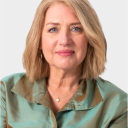 A middle-aged woman with shoulder-length blonde hair wearing a light teal satin blouse, looking at the camera against a plain background.
