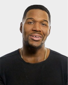 A man with short hair, a trimmed beard, and a black shirt is facing the camera against a plain light background.