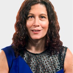 A woman with wavy brown hair wearing a sleeveless blue top with a black geometric mesh panel, posing against a plain light background.