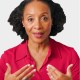 A woman with curly hair wearing a red collared shirt gestures with both hands while looking forward.