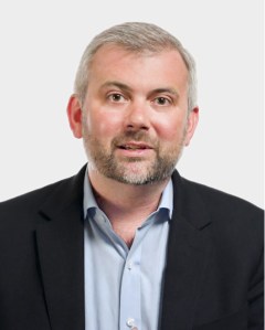 A man with short gray hair and a beard, wearing a dark blazer over a light blue shirt, poses against a plain light background.