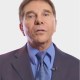 A man in a blue suit, tie, and collared shirt, wearing a lapel microphone, poses against a plain white background.