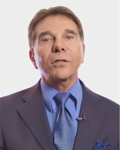 A man in a blue suit, tie, and collared shirt, wearing a lapel microphone, poses against a plain white background.