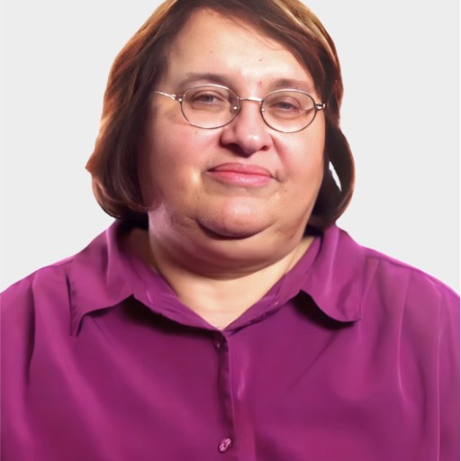 A middle-aged woman with short brown hair, glasses, and a purple button-up shirt is looking at the camera against a plain light background.