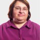 A middle-aged woman with short brown hair, glasses, and a purple button-up shirt is looking at the camera against a plain light background.